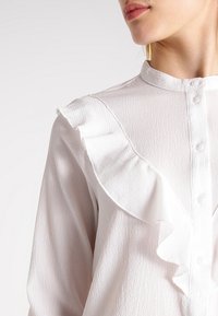 Blouse blanche texturée avec un col à volants, fermeture à boutons et manches longues. Le tissu présente un motif discret en relief.