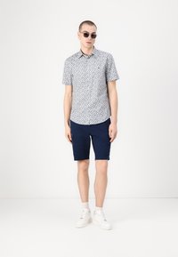 Kortermet skjorte med knappestolpe i marineblått blomstermønster, kombinert med marineblå shorts og hvite sneakers. Modellen har på seg solbriller og et sølvarmbånd.