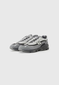 Le sneakers sportive presentano una tomaia sintetica grigia con accenti in rete, una suola nera spessa e dettagli riflettenti. Numero di stile '1000' visibile.