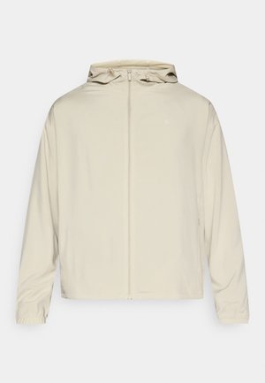 Casaco bege claro com fecho de correr frontal, capuz ajustável e punhos elásticos. Textura lisa com forro em rede. Design minimalista com detalhe de logotipo.