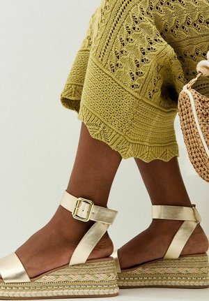 Dunkelhäutige Beine in hellgoldfarbenen Keilsandalen mit Knöchelriemen, kombiniert mit einem strukturierten olivgrünen Strickrock und einer gewebten Strohtasche.