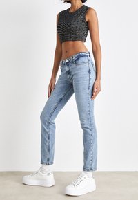 Kvinna som bär en svart croppad topp med logotyptext, ljusblå högmidjade jeans, vita strumpor och vita platåskor, stående på sidan.