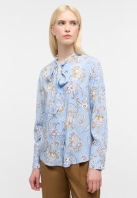 Blusa azzurro chiaro con motivo paisley in beige e marrone, caratterizzata da un collo legato, maniche lunghe e una vestibilità leggermente ampia. Tessuto morbido.