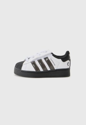 Biało-czarne buty sportowe Adidas Superstar z czarnymi paskami, czarnym czubkiem i podeszwą oraz paskiem na rzep na jasnoszarym tle.