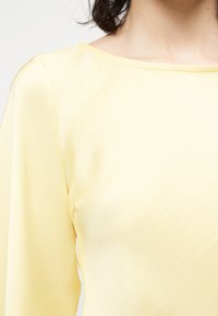 Žena oblečená v bledozltom tričku s dlhým rukávom a širokým okrúhlym výstrihom, zobrazená od ramien po stred trupu.