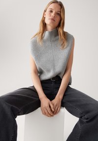 Junge Frau mit langen blonden Haaren, die einen grauen ärmellosen Rollkragenpullover und schwarze weit geschnittene Jeans trägt, sitzt mit gefalteten Händen.