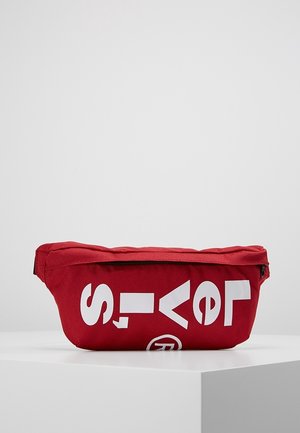 Sac banane en tissu rouge avec des lettres blanches en gras partiellement visibles à l'avant, posé sur une surface blanche sur un fond clair uni.