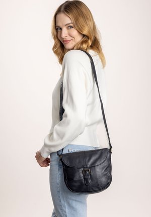 Schwarze Leder-Crossbody-Tasche mit gewölbtem Klappenverschluss, Schnallenakzent und verstellbarem Tragegurt, getragen von einer Person in einem hellen Pullover und Jeans.