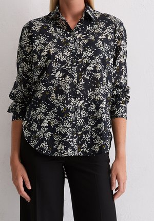 Vrouw draagt een zwart overhemd met knopen met witte en gele bloemenmotieven en lange mouwen, gecombineerd met een zwarte broek.