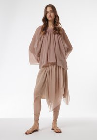 Blusa de gasa rosa claro con un corte holgado y escote fruncido, combinada con una falda beige con capas. Sandalias planas sencillas con tiras.