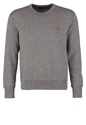 Mies, jolla on harmaa pitkähihainen paita, tummansiniset farkut ja mustat lenkkarit, seisoo yksinkertaisen valkoisen taustan edessä.