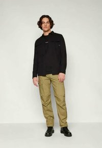 Μαύρο πουκάμισο με κουμπιά, σκούρο μπλουζάκι από κάτω, καφέ cargo παντελόνι και μαύρες μπότες φορεμένα από ένα μοντέλο σε ουδέτερο φόντο.