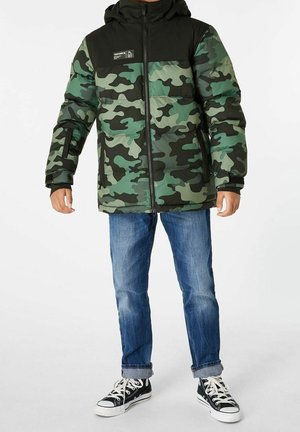 Dziecko w zielono-czarnej kamizelce puchowej w kamuflaż, niebieskie podwinięte dżinsy oraz czarne trampki za kostkę z białymi sznurowadłami.