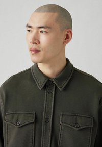 Camisa de color verde oscuro con botones, cuello con solapa, dos bolsillos delanteros y tejido texturizado. Presenta detalles de costura sutiles.