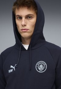Marineblå hættetrøje lavet af blødt stof, med et stort grafisk logo og accentbrand i hvid. Inkluderer en hætte med snører og raglanærmer.