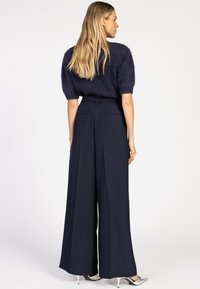 Blonde vrouw in een marineblauwe mohair trui met korte mouwen, hoge taille en een wijde pijp in marineblauw, met zilveren puntige hakken, zittend met de zijkant naar voren.