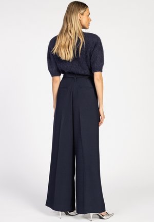 Blonde vrouw in een marineblauwe mohair trui met korte mouwen, hoge taille en een wijde pijp in marineblauw, met zilveren puntige hakken, zittend met de zijkant naar voren.