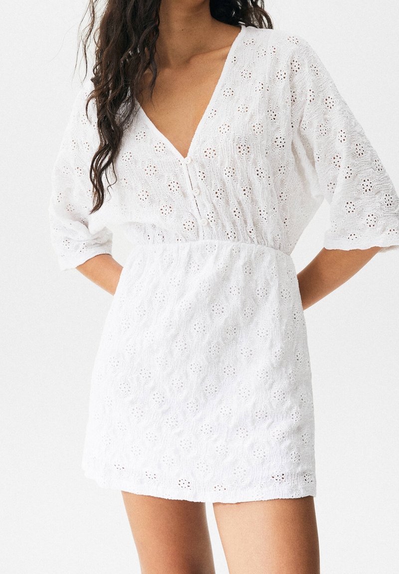 Femme portant une robe blanche en dentelle ajourée avec des manches jusqu'au coude et un profond décolleté en V boutonné devant, debout les mains sur les hanches.