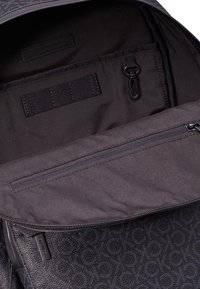 Interno dello zaino nero con tessuto liscio, tasca con zip e etichetta di branding. Presenta un esterno testurizzato con un motivo a logo ripetuto.
