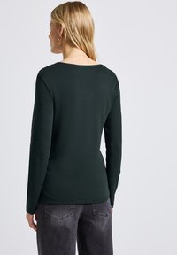 Tmavě zelená košile s dlouhým rukávem, pohodlného střihu a kulatým výstřihem. Hladká textura, nošena s šedými kalhotami, ukazuje jednoduchý design.