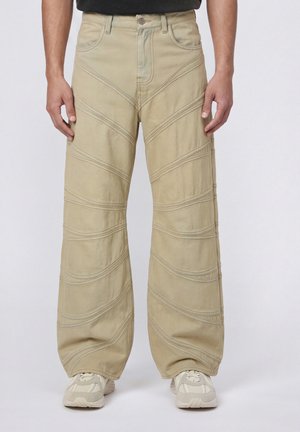 Pantalon beige à jambes larges avec des motifs cousus courbés, porté avec des baskets de couleur claire et un haut noir, visible de la taille aux pieds.