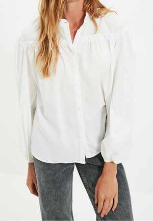 Femme portant un chemisier blanc à manches longues avec épaules froncées et poignets larges, associé à un jean denim gris foncé.
