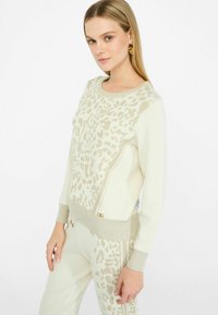 Maglione crema con motivo leopardato beige, polsini e orlo a coste. Presenta una cerniera laterale e un accento dorato decorativo sull'orlo.