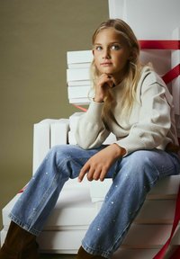 Maglione beige chiaro, jeans di denim blu con decorazioni in argento e stivali marroni contro scatole bianche con nastri rossi. Stile morbido e casual.