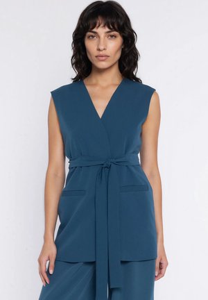 Kobieta o ciemnych włosach do ramion, ubrana w bezrękawnik turkusowy top z wiązanym paskiem i pasujące spodnie, stojąca na tle białego tła.