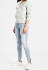 Sudadera gris con un logo en rojo. Vaqueros ajustados azul claro con desgaste y aberturas laterales. Zapatillas blancas completan el conjunto.