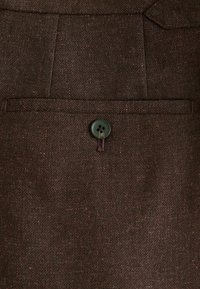 Tissu brun avec une texture subtile, comportant une poche poitrine unique et un bouton noir à deux trous. Design ajusté.