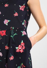 Abito floreale in tessuto blu scuro, con fiori e foglie colorati. Il design include una gonna svasata e dettagli con tasche laterali.