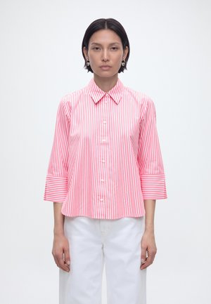 Kobieta z krótkimi ciemnymi włosami, ubrana w różowo-białą koszulę zapinaną na guziki z pionowymi paskami i rękawami 3/4 oraz białe spodnie na białym tle.