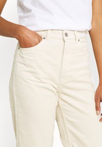 Cremfarbene Cordhosen mit hoher Taille, versehen mit Fronttaschen und subtiler vertikaler Rippstruktur, Knopfverschluss und Gürtelschlaufen.