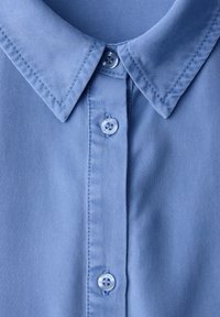 Chemise bleu clair à col avec trois boutons assortis visibles, gros plan montrant les détails de la couture sur le col et la patte de boutonnage.