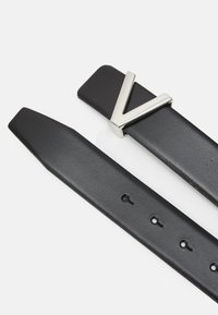 Čierny kožený opasok s kovovou prackou v štýlovom tvare "V". Pás je hladký s rovnomerne rozmiestnenými dierkami na nastavovanie.