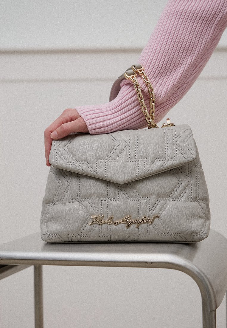 Eine Hand hält eine hellgraue, gesteppten Handtasche mit goldener Kette und Signatur, die auf einem Metallstuhl ruht, während ein rosa, geripptes Sweatshirt getragen wird.