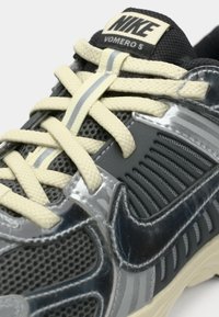Αθλητικό παπούτσι Nike Vomero 5 σε μαύρο και γκρι χρώμα με κρέμ κορδόνια και επάνω μέρος από δίχτυ, με λεπτομερή πλάγια και πάνω όψη.