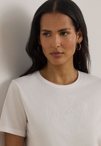 Γυναίκα με μακριά σκούρα μαλλιά που φοράει χρυσούς κρίκους στα σκουλαρίκια και λευκό T-shirt με ένα διακριτικό ανάγλυφο κυκλικό λογότυπο στο στήθος.