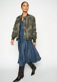 Maslinasto zelena bomber jakna s patentnim zatvaračima i rebrastim manžetama preko lepršave plave haljine, uparena sa sjajnim crnim čizmama do koljena.