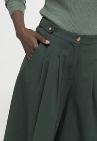 Donkergroene plooibroek met een hoge tailleband met twee knopen en zijzakken. Gemaakt van een soepele stof met een subtiele textuur.