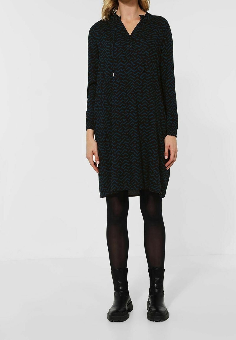 Femme portant une robe longue à manches longues sombre à motifs, des collants noirs et des bottines noires, debout devant un fond uni.