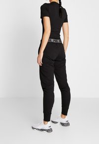 Kobieta z warkoczami, ubrana w czarny dopasowany t-shirt, czarne dresowe spodnie z markowym paskiem oraz białe trampki z czarną, wzorzystą podeszwą.