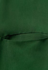 Tissu vert foncé avec une texture lisse, présentant une simple poche cachée le long de la couture. Pas de quincaillerie ou de motifs visibles.