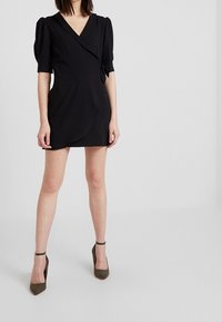 Black wrap dress with short puff sleeves, V-neckline, and a side tie detail. Worn with olive green heeled shoes. Smooth fabric.