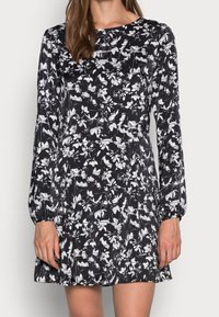 Svart klänning med långa ärmar, prydd med ett vitt blommönster. Gjord av tyg med en slät textur, något utsvängd nederst.