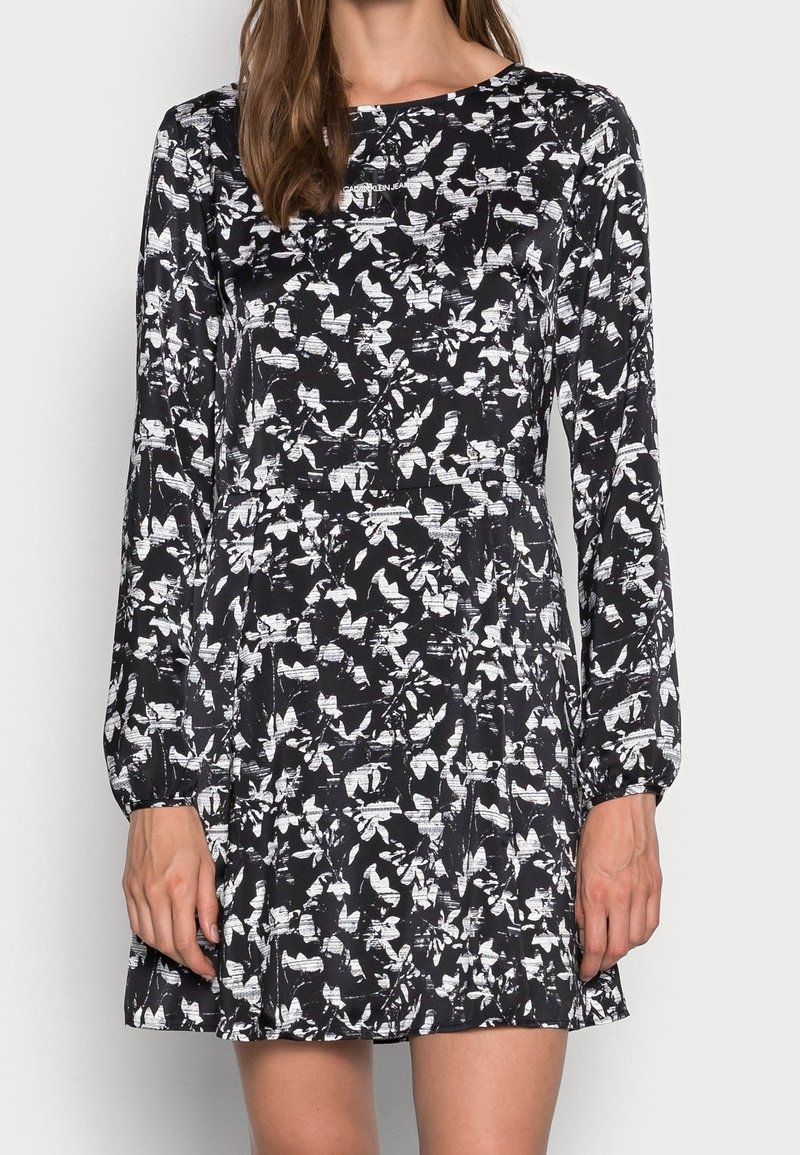 Svart klänning med långa ärmar, prydd med ett vitt blommönster. Gjord av tyg med en slät textur, något utsvängd nederst.