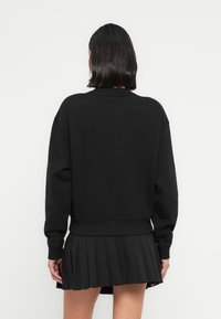 Personne aux cheveux courts et foncés portant un sweat-shirt noir oversize et une jupe plissée noire, debout dos à un fond uni.