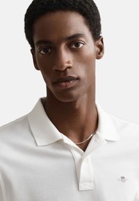 Valkoinen poolopaita, jossa on klassinen kaulus ja pieni kirjailtu logo. Sileä pinta, pehmeä kangas ja vakiomallinen leikkaus.