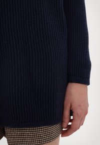 Sini ribbed knit neule, jossa on tekstuuripinta ja pitkät hihat, yhdistettynä ruskeaan haukansilmää kuviolliseen hameeseen, joka näkyy alareunasta.